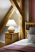 Ceramic lamp on bedside cabinet and wooden bed below sloping ceiling with rustic wooden beams