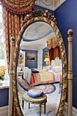 Oval cheval mirror with carved frame reflecting antique footstool and luxurious double bed with half-tester canopy