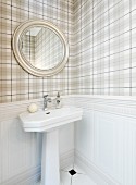 Oval mirror above pedestal sink and pale tartan wallpaper above white wainscoting