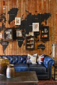 Vintage, blue leather couch against wooden wall with painted world map, small wall-mounted cabinets and pictures; rustic containers on coffee table in foreground