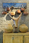 Faux animal skull amongst woven vessels on pale wooden cabinet below hand-crafted cow bells hanging from hooks on clay wall