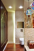 Bunte Kugelleuchten über Waschtisch mit Kiesel-Mosaikfliesen, dahinterliegendes WC-Abteil mit indirekter Beleuchtung; seitlich ein eingebauter Wandschrank