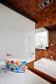Modern bathroom with areas of wall and ceiling clad in a mosaic of wooden blocks, integrated sink and corner bathtub with decorative floral pattern on front
