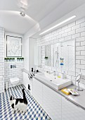 Cat in white, modern bathroom with tiled walls & floor, long washstand with base cabinets & strip light over mirror; modern artwork above wall-mounted toilet in background