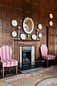 Open fireplace flanked by white and pink striped chairs, decorative china wall plates on wood-panelled wall and hand-made Aubusson-style rug