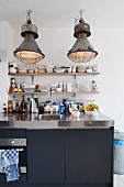 Vintage, industrial-style pendant lamps above modern kitchen counter with stainless steel worksurface and black doors; minimalist shelves of crockery on wall