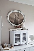 Small glass-fronted cabinet on chest of drawers below religious painting in oval frame on pastel grey wall