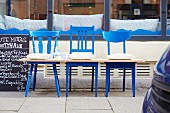 Drei blaue Holzstühle mit Holzplatte zu einer Sitzbank umfunktioniert vor Restaurant
