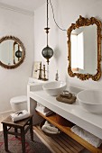 Rustic wooden stool in front of white washstand with twin basins below vintage mirror with ornate gilt frame