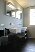 Black, curved washstand with white countertop basins and wall-mounted taps below illuminated mirrors on grey-painted wall