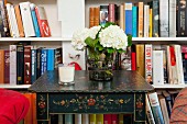 Vase of hydrangeas on side table with floral, country-style painting in front of fitted bookcases