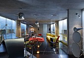 Metal counter, bar stools and yellow sofa on black stone floor below chrome lights suspended from exposed concrete ceiling in open-plan, modern interior