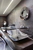 Two curved chrome basins on solid wooden washstand against stone-tiled wall