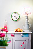 Wall clock decorated with colourful fabrics and neon green pompoms, vintage sideboard and table lamp made from stacked porcelain crockery
