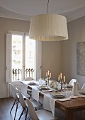 Classic pendant lamp with fabric lampshade above white place settings on wooden dining table with classic chairs
