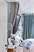 Half-tester bed with many scatter cushions and blue and grey striped canopy against floral wallpaper in romantic bedroom in shades of blue