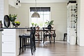 Black bar stools at white counter and dining area below window in open-plan kitchen with white wood cladding