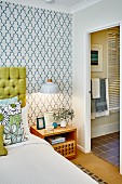 Illuminated bedside table below pendant lamp against retro patterned wallpaper; door leading to ensuite bathroom to one side