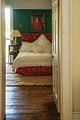 View through open door into bedroom with wooden floor and lace bedspread on red bed