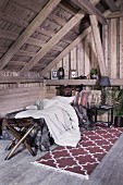 Bett mit heller Decke, davor Hocker mit Fellbezug und gemusterter Teppichläufer, in rustikalem Dachraum mit Holzkonstruktion