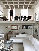 Open-plan interior with white corner sofa, woman on gallery with metal balustrade and dining area below concrete coffered ceiling