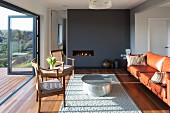 Antique chair and leather sofa around round coffee table on rug in front of fireplace in dark grey partition wall; open folding terrace doors with view of wooded landscape
