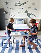 Zwei Kinder spielen im Kinderzimmer am Tisch auf blauweiß gestreiftem Teppich, im Hintergrund verschiedene Wandtattoos mit Flugzeugmotiven