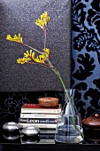 Stacked books, tealight holders and yellow Anigozanthos in glass vase on surface against wall covered in ornate wallpaper