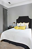 Double bed with dark headboard and scatter cushions arranged on white bedspread in bedroom with walls painted paint grey and traditional ambiance