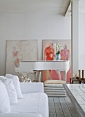 Sofa with white loose cover and grand piano in open-plan living area with modern paintings leaning against whitewashed brick wall