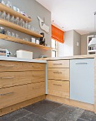Corner of kitchen counter with solid wooden drawers below glasses on wooden shelves mounted on wall painted pale grey