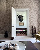 Coffee table in traditional living room with ornate wallpaper and view through open door of water buffalo hunting trophy on wall