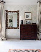 Antique wall mirror with elaborate frame next to dark wooden chest of drawers against ornately patterned wallpaper