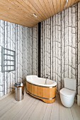 Wooden bathtub and toilet in corner of modern bathroom with tree-patterned wallpaper