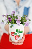 Woman's hands holding viola planted in white, painted mug