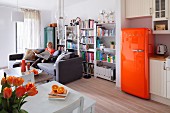 Open-plan living area with orange retro fridge; woman sitting on couch in front of bookcase