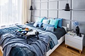Breakfast tray on double bed with scatter cushions and blankets in shades of blue; pale grey back wall with panelled structure