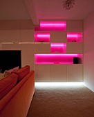 Fitted cabinets with shelf compartments illuminated bright pink behind orange sofa in dark interior