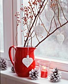 Branches of miniature rose hips and china love-hearts in red jug on windowsill