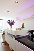 Island counter with white, polished surface and integrated, pale purple LED lighting in open-plan kitchen