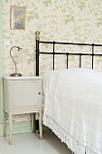 Ländliches Nachtkästchen neben Bett mit schwarzem, schmiedeeisernem Gestell, vor tapezierter Wand mit floralem Muster