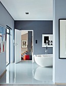 Blick durch Durchgang auf glänzenden Fliesenboden in blau-grauem Bad mit freistehender weißer Badewanne