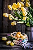Gelber Tulpenstrauß in Krugvase auf rustikaler Holzunterlage mit pastellfarbenem Flechtkörbchen und Ostereiern