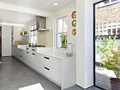 White, designer kitchen with raised hob beneath extractor hood, grey-tiled floor and floor-to-ceiling window with view of neighbouring building