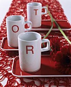 Three white china mugs printed with red letters and gerbera daisies on red trays on red, plastic table runner