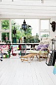 Cushions and fur rug on comfortable wicker lounger on white wooden floor in front of window with storage boxes on fitted shelving below sill
