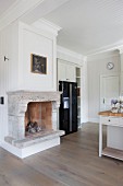 Open fireplace with stone surround next to modern, black fridge-freezer in white, wood-clad, country-house kitchen