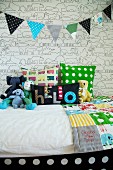 Colourful bedspread and soft toys on bed below bunting against wallpaper with graphical pattern of animals