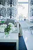 Vase of white peonies on coffee table and sofa in front of curtains with large black and white floral pattern