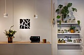 Counter with recessed wooden worksurface below dainty pendant lamps next to foliage plants on wall-mounted shelves